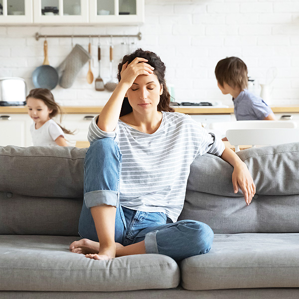 Young tired single mother suffers from headache closed eyes touch forehead sitting on couch while her daughter and son running around her and shouting, female babysitter feels exhausted by noisy kids